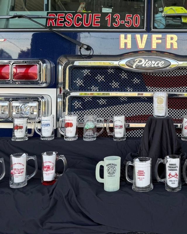 Carnival glasses lined up on black cloth in front of Rescue 13-50's flag grille: Hegins Fire Co. glasses on the left, a green Hegins Valley Fire-Rescue glass and this year's glass raised on a stand.